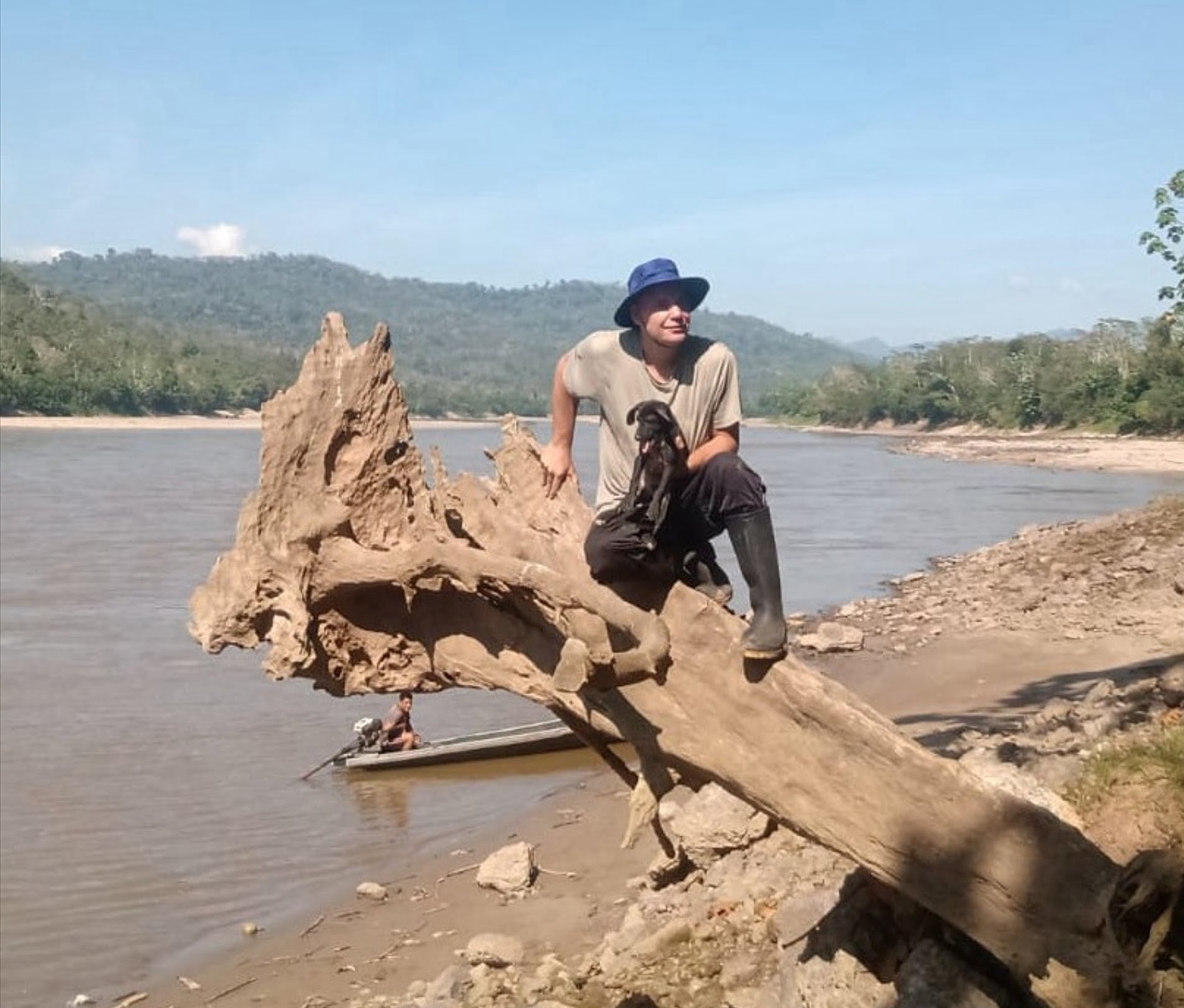 An image of Melvin Lok is sitting on a big tree on the beach of the Huallaga River. He's holding a dog, and there's a man in a boat in the background.